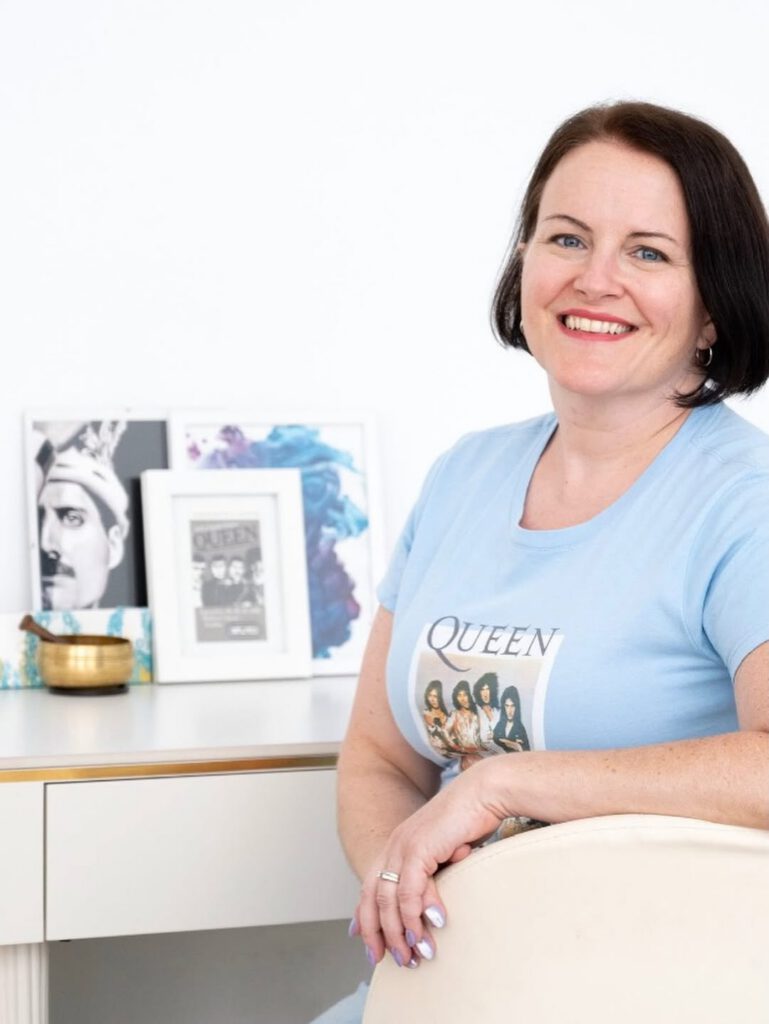 Sarah leans on the back of a cream armchair in a Queen T-shirt, smiling. Framed Freddie Mercury and Queen prints and a brass singing bowl sit on the desk behind her.