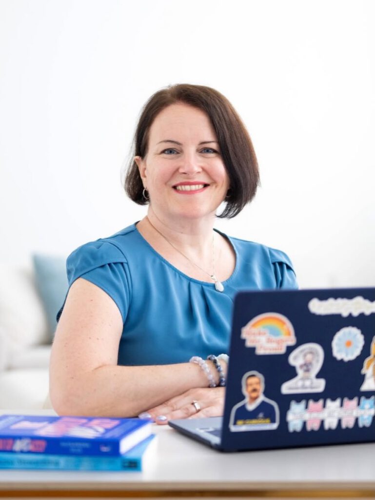 Sarah smiles at the camera from behind an open laptop covered in stickers, including a rainbow, a row of cats in trans flag colors, and a Ted Lasso sticker. A stack of books sits beside her.