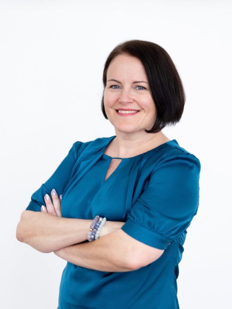 Sarah, a white woman with a chin-length dark bob and blue eyes, smiles with her arms folded. She wears a teal blouse and a few beaded bracelets in lavender, blueish purple, and sky blue.