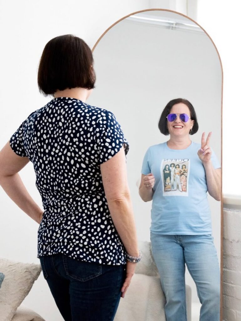 Sarah stands with her back to the camera in a navy polka-dot top, facing a tall arched mirror. Her reflection grins back in purple mirrored sunglasses and a Queen T-shirt, throwing a peace sign.