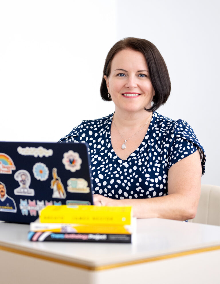 Sarah smiles at the camera from behind a laptop covered in stickers: a rainbow, a row of cats in trans flag colors, and Freddie Mercury mid fist-pump in his yellow jacket. Three books are stacked on the desk in front of her.
