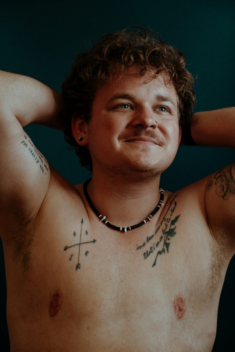 A man with tattoos across his chest and arms and faint top surgery scars. He's smiling at something out of frame.